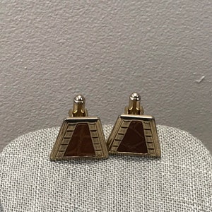 Puede incluir: Un par de gemelos de oro con una incrustaci&oacute;n de imitaci&oacute;n de cuero marr&oacute;n. Los gemelos tienen forma de trapecio.