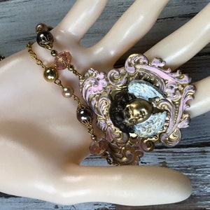 May include: A vintage-style necklace featuring a pendant with a cherub design. The pendant has a gold-colored cherub face with white wings, set against a pink and gold filigree frame. The necklace includes gold-colored chain and beaded accents.