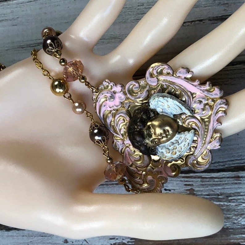 May include: A vintage-style necklace featuring a pendant with a cherub design. The pendant has a gold-colored cherub face with white wings, set against a pink and gold filigree frame. The necklace includes gold-colored chain and beaded accents.