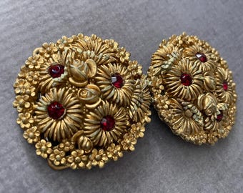 May include: Two circular gold-toned metal brooches featuring a dense, textured floral design. Each piece is adorned with small, raised flower petals and multiple deep red, round faceted stones embedded within the center of the floral clusters.