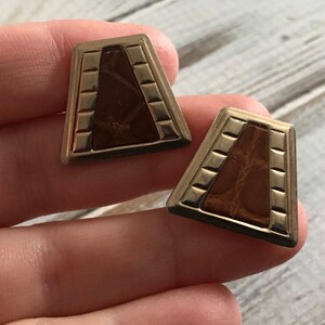 Puede incluir: Un par de pendientes de tono dorado con un dise&ntilde;o geom&eacute;trico. Los pendientes presentan una incrustaci&oacute;n de imitaci&oacute;n de cuero marr&oacute;n.