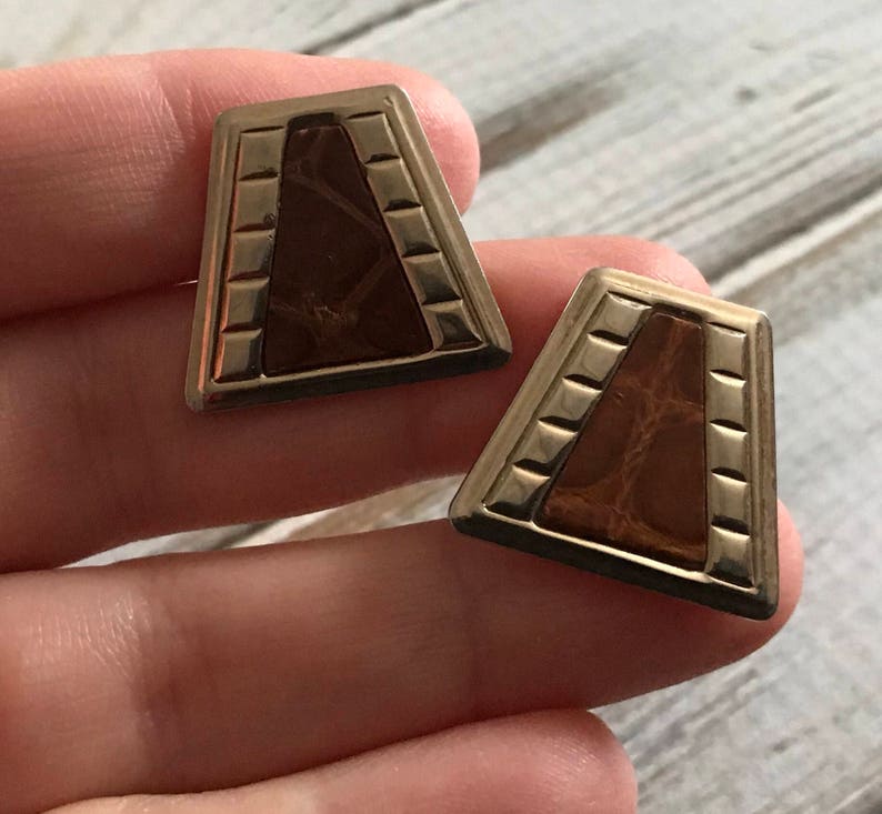 Puede incluir: Un par de pendientes de tono dorado con un dise&ntilde;o geom&eacute;trico. Los pendientes presentan una incrustaci&oacute;n de imitaci&oacute;n de cuero marr&oacute;n.