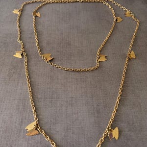 May include: Gold-coloured necklace with a delicate chain design. The necklace is decorated with small butterfly charms. The jewellery is displayed against a neutral grey background, highlighting the warm metallic colour of the necklace.