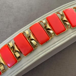 May include: A vintage bracelet featuring rectangular coral-red panels set in gold-tone metal. The bracelet has a series of connected links, with each panel framed by ornate, curved metalwork. The bracelet is displayed on a white surface.