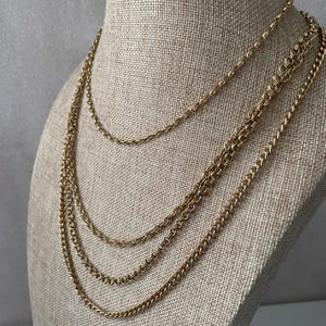 May include: A layered gold-tone necklace set consisting of three distinct chains. The top chain is a delicate cable link, while the middle and bottom chains feature thicker, interlocking curb links, all displaying a polished metallic finish.