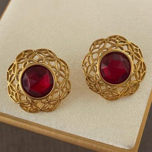 May include: A pair of gold-toned stud earrings. Each earring features a large, round, faceted, deep red gemstone set within an openwork, floral-inspired frame. The earrings are displayed on a cream-colored surface.