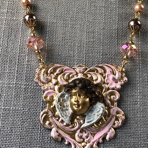 May include: A vintage-style necklace featuring a pendant with a cherub face and wings, painted in gold, pink, and white. The pendant is suspended from a chain with pink and brown beads. The necklace is a unique piece of jewelry.
