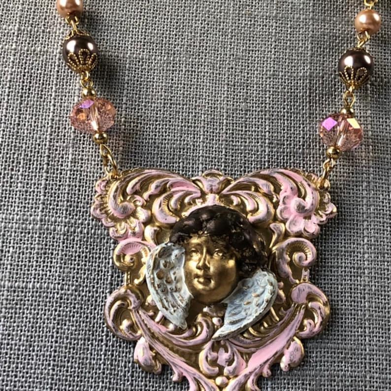 May include: A vintage-style necklace featuring a pendant with a cherub face and wings, painted in gold, pink, and white. The pendant is suspended from a chain with pink and brown beads. The necklace is a unique piece of jewelry.