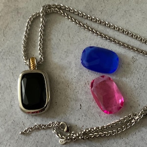 May include: A silver-tone chain necklace featuring a rectangular pendant with a faceted black stone and a gold-tone accent at the top. Two additional loose faceted stones, one blue and one pink, are positioned next to the pendant necklace.