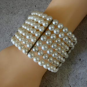 May include: A multi-strand bracelet featuring seven rows of round, cream-colored faux pearls. The strands are held together by a vertical, thin, dark-toned metal bar that spans the width of the bracelet, creating a structured, layered appearance.