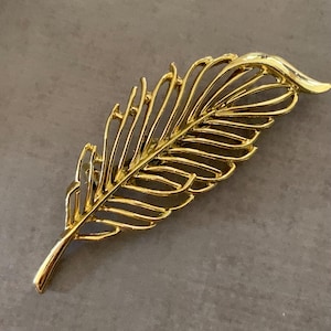 May include: A gold-colored feather-shaped brooch. The brooch has a detailed openwork design, with multiple parallel lines creating the feather's structure. The pin is made of a shiny, reflective metal, and is set against a neutral background.