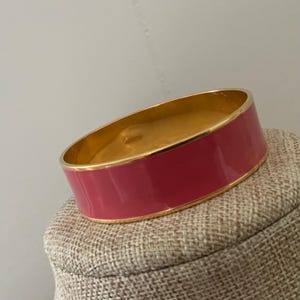 May include: A gold-tone bangle bracelet with a vibrant pink enamel band. The bracelet has a smooth, polished finish and is displayed on a neutral-toned surface. This piece of jewelry is a stylish accessory.