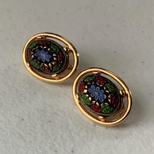 May include: A pair of gold-toned oval earrings. Each earring features a central design with a black background, adorned with a floral pattern in blue, red, and green. The earrings have a decorative gold frame.