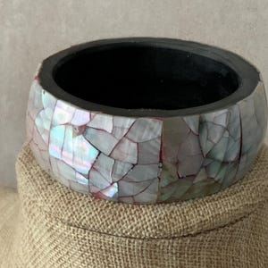 May include: A wide bangle bracelet featuring a mosaic of iridescent mother-of-pearl shell fragments in shades of white, pink, and blue. The interior of the circular bracelet is finished in a solid matte black, providing a contrast to the shell.