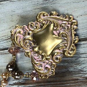 May include: A vintage-style, ornate brooch with a gold-colored base and pink accents. The brooch features a heart-shaped design with intricate scrollwork and is adorned with a beaded chain of brown and pink beads.