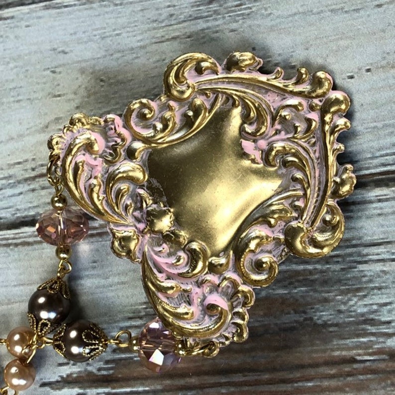 May include: A vintage-style, ornate brooch with a gold-colored base and pink accents. The brooch features a heart-shaped design with intricate scrollwork and is adorned with a beaded chain of brown and pink beads.