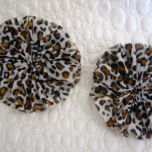 May include: Two round, ruffled fabric pieces with a leopard print pattern. The print features black and brown spots on a white background. The fabric is gathered in the center, creating a flower-like shape. The items are on a white, textured surface.