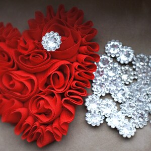 May include: A red fabric heart-shaped decoration with a large rhinestone in the center. The heart is surrounded by a cluster of smaller rhinestones.