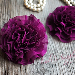 May include: Two large, deep purple fabric flowers with ruffled petals. The flowers are on a wood background.