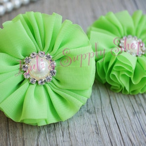 Op de afbeelding: Twee limoengroene stoffen bloemen met gerimpelde bloemblaadjes en een parel- en strass-middenstuk. De bloemen liggen op een houten oppervlak met een parelsnoer op de achtergrond. Waarschijnlijk voor knutselen of versieringen.