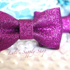 May include: Two magenta glitter bow hair clips. The bows are made of a sparkly fabric and are tied in a classic bow shape. The bows are sitting on a white surface with white pearls.