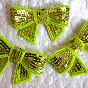 May include: Three lime green fabric bows with gold sequins. The bows are sewn with a decorative stitch around the edges.