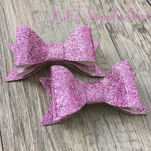May include: Two pink glitter hair bows. The bows are made of a sparkly pink material and are tied in a classic bow shape.