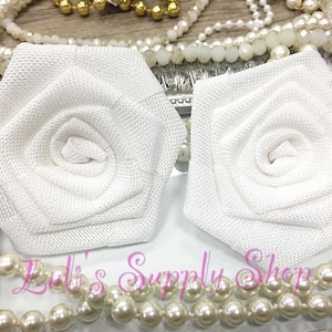 May include: Two white fabric rose flower embellishments. The roses are made of a textured fabric and have a simple, elegant design.