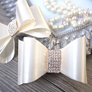 May include: Two ivory satin hair bows with rhinestone embellishments. The bows are tied in a classic bow shape and have a rectangular rhinestone centerpiece. The bows are displayed on a wooden surface.