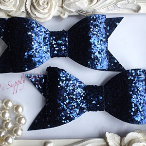 May include: Two navy blue glitter hair bows with a light blue shimmer. The bows are displayed on a white background with white roses.