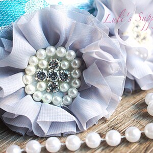 May include: A gray fabric flower with a pearl and rhinestone center. The flower is on a wooden surface with a pearl necklace.