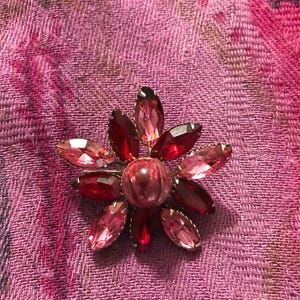 May include: A red and pink flower-shaped brooch with a large, round, red center stone. The brooch is made of rhinestones and has a gold-tone setting.