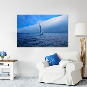 May include: A canvas print of a sailboat on the ocean under a blue sky with a large cloud formation. The artwork is displayed above a white armchair with a blue pillow, a side table, and a floor lamp.