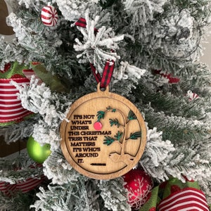 May include: A round wooden Christmas ornament with a green tree and red ornament. The ornament has the text "It's not what's under the Christmas tree that matters. It's who's around it."