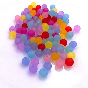 Op de afbeelding: Een verzameling kleurrijke, ronde kralen in verschillende tinten rood, geel, oranje, roze, paars en blauw. De kralen hebben een matte afwerking en zijn verspreid over een wit oppervlak. Geschikt voor het maken van sieraden en knutselen.
