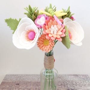 Peut inclure: Un bouquet de fleurs en feutre dans un vase en verre transparent. Les fleurs sont blanches, roses et orange. Le vase est enveloppé de ficelle.