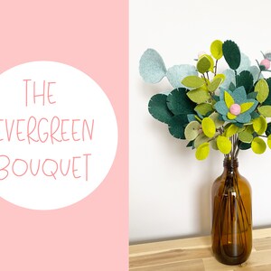 K&ouml;nnte beinhalten: Ein Blumenstrau&szlig; aus Filz in Gr&uuml;n- und Rosat&ouml;nen, arrangiert in einer braunen Glasvase. Der Strau&szlig; ist mit "The Evergreen Bouquet" beschriftet.