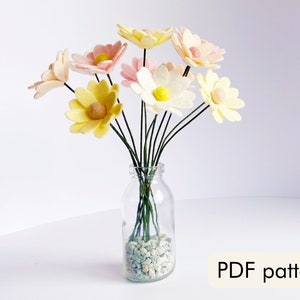 May include: A bouquet of felt flowers in shades of yellow, pink, and white, arranged in a clear glass vase with small white stones at the bottom. The vase is on a white surface. PDF pattern.