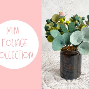 K&ouml;nnte beinhalten: Eine Sammlung von Filzlaub in einer braunen Glasvase. Das Laub ist in Gr&uuml;nt&ouml;nen gehalten und enth&auml;lt kleine rosa Filzblumen. Der Text "MINI FOLIAGE COLLECTION" befindet sich auf einem rosa Hintergrund.