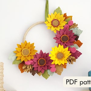 May include: A felt sunflower wreath with burgundy and yellow flowers, green leaves, and brown accents. The wreath is hanging on a white wall. PDF pattern.
