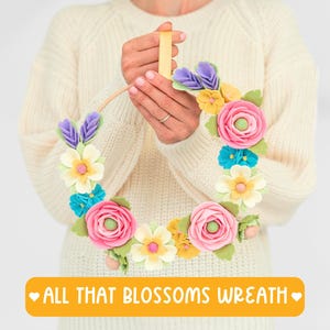 May include: A colorful floral wreath with pink, yellow, blue, and purple felt flowers and green leaves. The wreath is held by a person wearing a cream-colored sweater. The text "ALL THAT BLOSSOMS WREATH" is on a yellow banner.