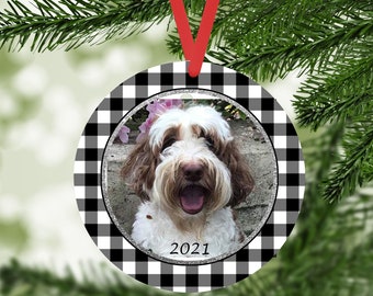 May include: A round, white and black checkered Christmas ornament with a photo of a dog inside. The photo is surrounded by a silver glitter border. The year 2021 is written in silver glitter at the bottom of the photo.