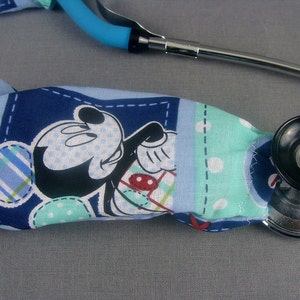 May include: A blue and white stethoscope cover with a Mickey Mouse design. The cover is made of fabric and has a patchwork design. The stethoscope is silver and black.