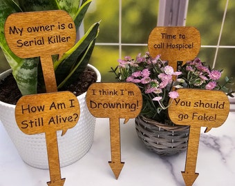 May include: Wooden plant markers with humorous phrases like "My owner is a Serial Killer" and "How Am I Still Alive?" are displayed in potted plants. The markers have a speech bubble shape and a pointed end for insertion.