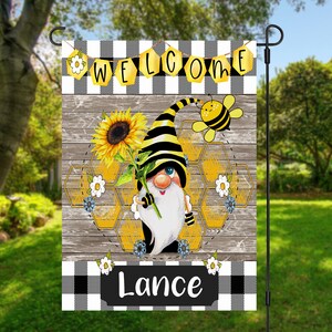 May include: A welcome garden flag with a black and white checkered background. A gnome with a black and yellow striped hat holds a sunflower. A bee flies above the gnome. The flag says "Welcome" and "Lance" at the bottom.