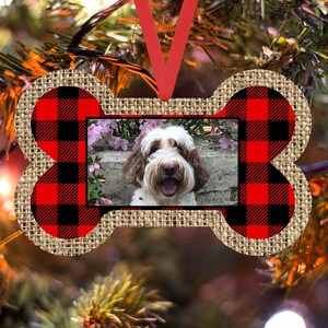 May include: A red and black buffalo plaid dog bone ornament with a photo insert featuring a brown and white dog. The ornament is made of burlap and has a red ribbon for hanging.