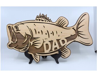 May include: A wooden bass fish cutout with the word "DAD" carved into the body. The fish is brown and light brown and is standing on two black metal stands.