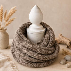 May include: A knitted infinity scarf in a warm brown hue. The chunky knit texture is visible, creating a cozy and stylish accessory. The scarf is draped around a white decorative object, suggesting a home decor or fashion theme.