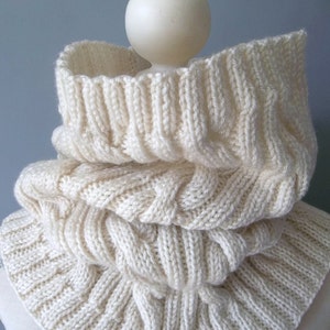 May include: A white knitted cowl neck scarf with a cable knit pattern.