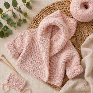 Puede incluir: Una chaqueta de bebé de punto rosa claro con capucha y puños enrollados. La chaqueta se exhibe sobre una estera tejida con una madeja de hilo a juego, agujas de tejer y una pequeña muestra de punto. La chaqueta está hecha de hilo suave y texturizado.